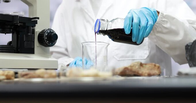 Food scientist in white lab coat and blue gloves pours dark liquid into a glass beaker beside a microscope and food samples in a laboratory.