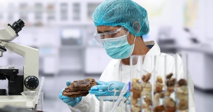 Masked laboratory scientist examines a soil sample with tools beside a microscope and test tubes in a bright research lab.
