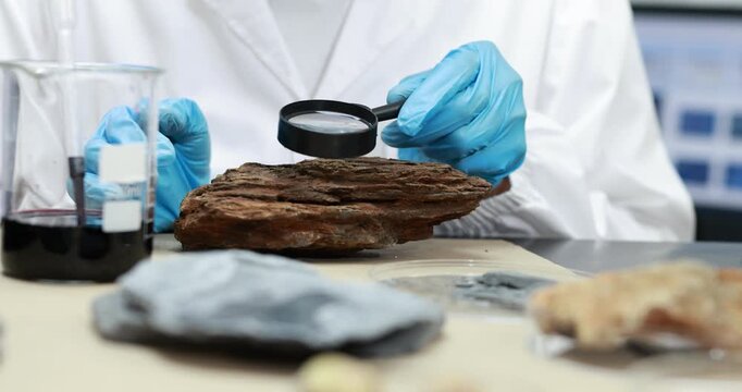 Scientist in a white coat and blue gloves examines layered rock samples with a magnifying glass in a laboratory, with beakers of liquid in the background.