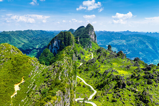 High angle aerial perspective shows a winding paved road traversing vibrant green karst mountain ridges under a bright sunny sky.