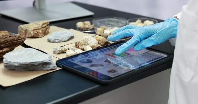 Scientist in blue gloves uses a tablet to examine and document rock samples on a laboratory workbench, combining field geology with digital research.