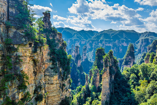 Tall sandstone pillar peaks rise above lush green forested valleys under bright blue sky with cumulus clouds in Zhangjiajie landscape.