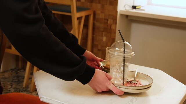 Barista lifts dirty dessert plate from small cafe table. Woman holds cup plate and cutlery together removing leftovers after sweet break