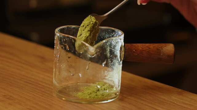 Barista scoops green matcha powder into glass pitcher on counter. Woman tips spoon carefully starting fresh green tea base for cafe drink
