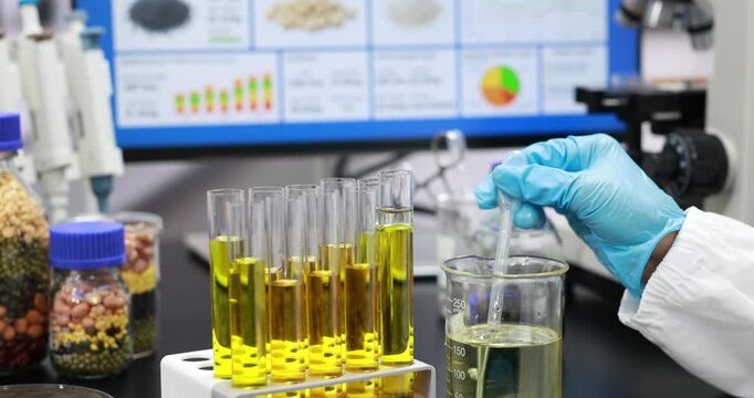 Laboratory technician in blue gloves handling glassware with yellow liquid samples, test tubes, beaker, microscope, and data charts in a modern research lab.