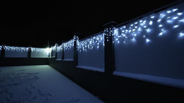 Snow blankets a dark, stone-walled pathway. Twinkling lights cascade along the fence, casting a cool glow. Frosty night adds quiet charm to the wintry scene. Perfect for holiday, winter