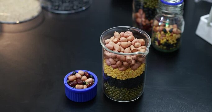 Glass jars and a blue lid display colorful layered dried beans, peas, lentils and grains on a dark table, with mixed legumes in the foreground and more jars in the background.
