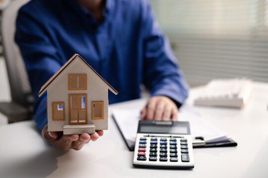 Professional holds a house model beside a calculator while reviewing mortgage documents. The scene represents home loan planning, property finance, affordability, budgeting, and real estate purchase.