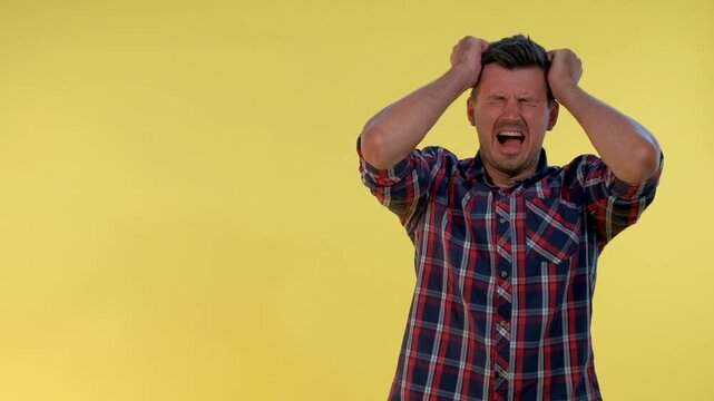 A young man wearing a red, blue, and white plaid shirt is holding his head in pain, simulating a severe headache against a solid yellow studio background.