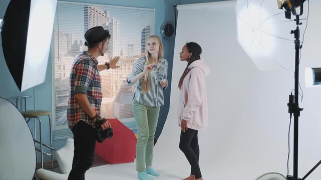 A male photographer directs two female models and a makeup artist in a professional indoor photography studio with lighting and urban cityscape backdrop.