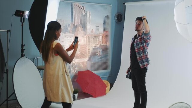 A young dark-skinned woman in a yellow dress takes photos with a smartphone while a light-skinned man in a plaid shirt holds a camera and gestures in a studio.