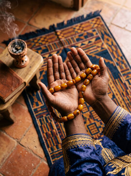 Overhead view of praying hands with amber beads over a West African textile, ideal for Islamic spirituality and cultural holiday campaigns.