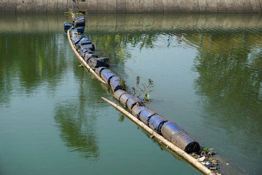 A makeshift floating trash barrier constructed from blue plastic barrels and bamboo floating on a river water surface to trap garbage. 