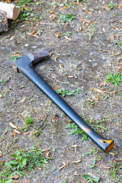 Axe Resting on the Ground for Firewood Preparation. A traditional axe with a dark metal head and long handle placed on the ground in a rural outdoor area. Sparse vegetation surrounds the tool, creatin