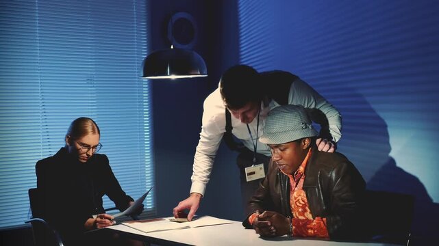 Three individuals examine documents around a table in an office interrogation room with dramatic lighting and horizontal blinds in the background.