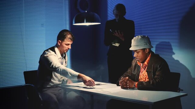 Three men in an interrogation room at nighttime review documents on a table under a dome-shaped lamp. Two are seated while a third stands behind with arms crossed.