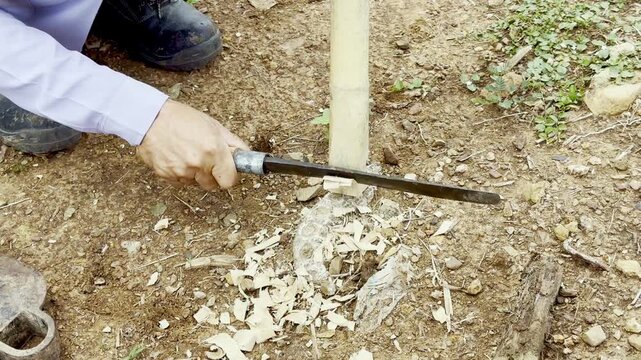 A Thai local craftsman uses a traditional Thai knife to carve bamboo for making a hoe handle, reflecting rural lifestyle and local craftsmanship.