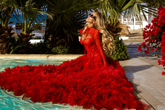 Stunning blonde model in elegant red tulle ballgown posing poolside with palm trees and flowers at a luxury tropical resort.