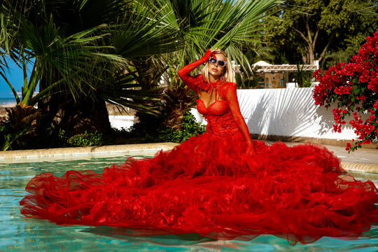 Stunning blonde model in elegant red tulle ballgown posing poolside with palm trees and flowers at a luxury tropical resort.