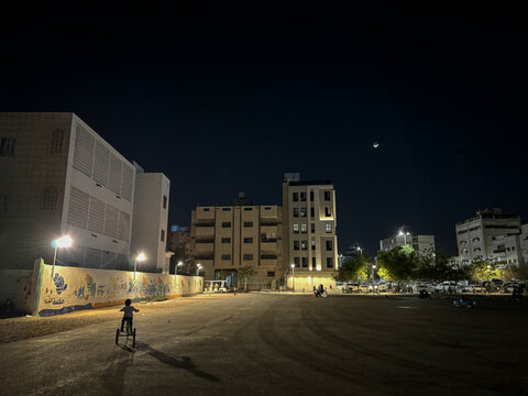 A child rides a tricycle across an empty courtyard beneath a crescent moon at night, Al Madinah Province, Madinah, Saudi Arabia