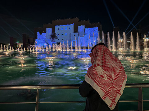A man in a keffiyeh watches a blue-lit fountain show in front of an illuminated heritage facade, Riyadh, Riyadh, Saudi Arabia