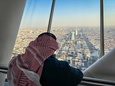 A visitor in a keffiyeh sits resting against the glass window of the Kingdom Centre sky bridge, Riyadh, Riyadh, Saudi Arabia
