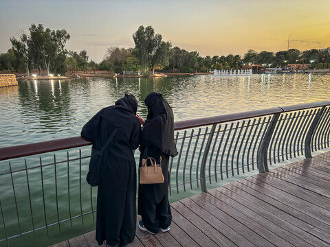 Two women in abaya watch the sunset over a park lake, Riyadh, Riyadh, Saudi Arabia