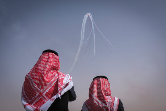 Two men in red and white headscarves look up at looping smoke trails left by aerobatic jets, Riyadh, Riyadh, Saudi Arabia