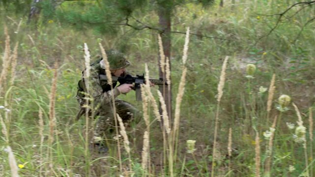 Soldier crouches low, concealed by tall grass. He holds rifle steady, eyes focused ahead. Dense forest shadows frame his camouflaged form. Natural light filters through green canopy above