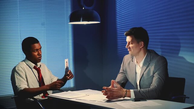 An African police detective shows photographs to a suspect during questioning in a professional office interrogation room with documents on the table.
