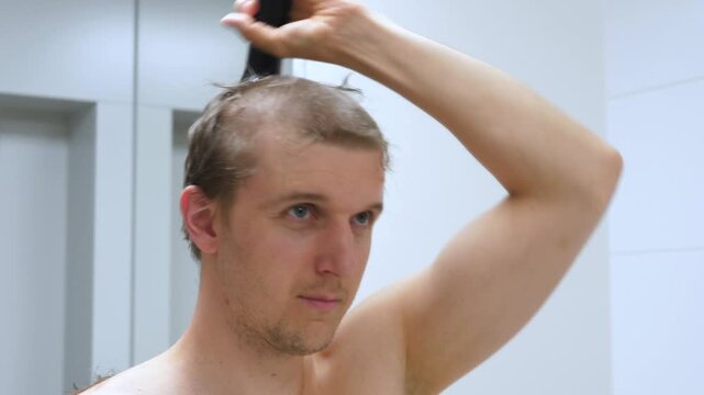 Man cuts his hair with electric clipper before mirror. Focused grooming continues in bathroom, with short hair being shaped carefully and bare shoulders visible under cool light as arm holds trimmer.