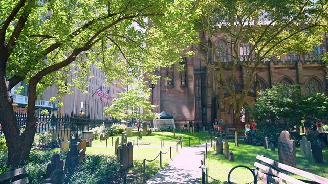 New York, USA, 1 August 2026 : Sunlit graveyard near Trinity Church in Lower Manhattan. Wooden bench and path border green grass field with ancient gravestones under leafy tree canopy.