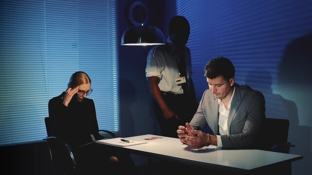 Three professionals engage in a serious discussion around a table in a modern office. One woman appears stressed while two men converse and observe the situation.
