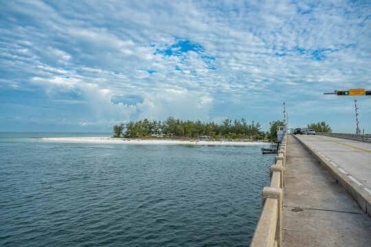 Concrete bridge structure overlooking sandy beach and green island. An elevated roadway features a pedestrian path viewing a coastal park shoreline and an ocean inlet under a blue sky.