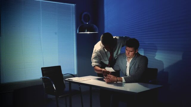 An African-descent detective leans over a seated Caucasian man during a tense nighttime interrogation in an office room with blinds and a desk lamp.