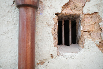 Fototapeta premium Old drainage pipe beside a barred ventilation opening on a cracked and weathered plaster wall, representing aging buildings, deterioration, maintenance, plumbing, renovation, and urban decay.