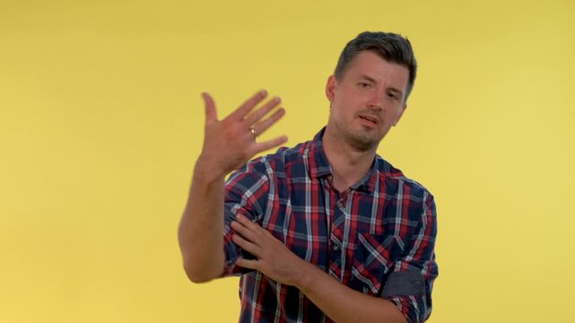 An adult man wearing a red, blue, and white plaid shirt shows disdain with crossed arms and then spreads his arms wide against a solid yellow indoor background.
