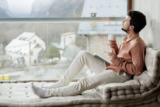 Man with coffee and notebook looking out through a large window at a village