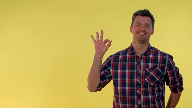 A young man wearing a red, blue, and white plaid shirt smiles and makes the okay hand sign in front of a solid yellow background in a studio setting.