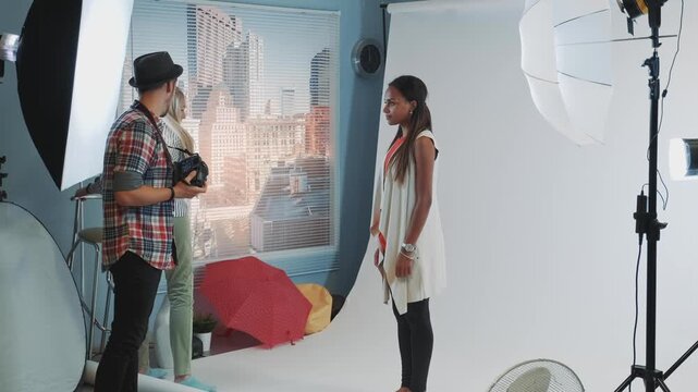 A male photographer takes pictures of two female models posing with a bar high chair in a professional indoor photography studio with urban cityscape backdrop.