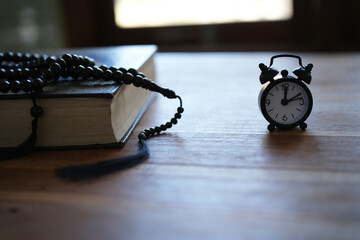 The Quran, clock, and prayer beads on a wooden table with dim lighting. Creates an atmosphere of spiritual contemplation and a sense of reverence. Reminds us of the time to pray. Muslim, Ramadan, Isla © PANCASONA CREATION