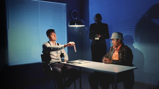 Two men sit at a table during an interrogation while a female detective stands taking notes in a police interview room with documents and blinds.