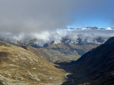 High-altitude panoramic mountain view in the Swiss Alps near Realp, Uri, showing jagged rocky peaks, dramatic ridges, alpine valleys and cloud layers under a deep blue sky in the wild landscape