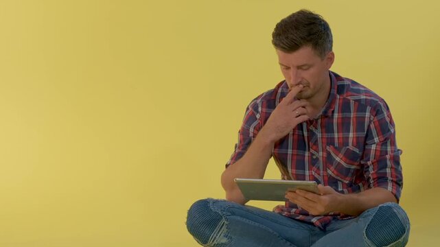 A young man wearing a red, blue, and white plaid shirt and blue jeans is seated and focused on using a digital tablet against a solid yellow background.