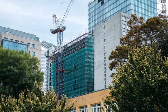 Building wrapped in scaffolding and green safety netting rises behind trees among mixed city blocks. Glass curtain wall installation, glazing panel lifted by crane, facade engineering