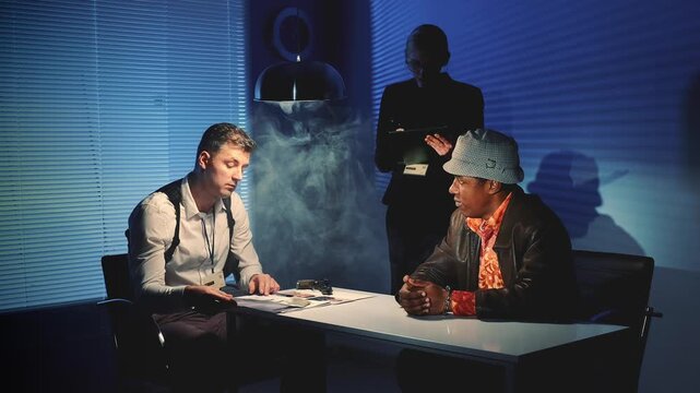 Three men in a nighttime interrogation room engage in a tense confrontation as one man points a handgun across the table while another gestures defensively.