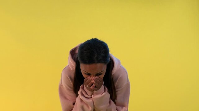 A young woman with long dark hair laughing and reacting emotionally while wearing a light pink hooded sweatshirt against a solid yellow background in a studio setting.