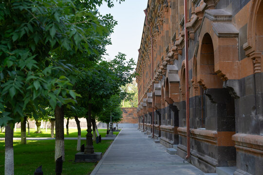 Quiet paved walkway lined with leafy trees beside an ornate historic stone building featuring arched windows and carved architectural details. T