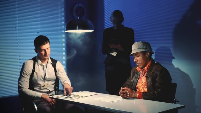 A detective and two men interact around a table in a nighttime office interrogation room with documents and blinds in the background.