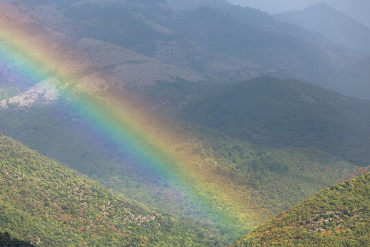 A bright rainbow arcs diagonally across lush green mountain ridges covered in soft mist. Beautiful atmospheric weather phenomena inspire profound feelings of hope, peace and deep spiritual renewal.